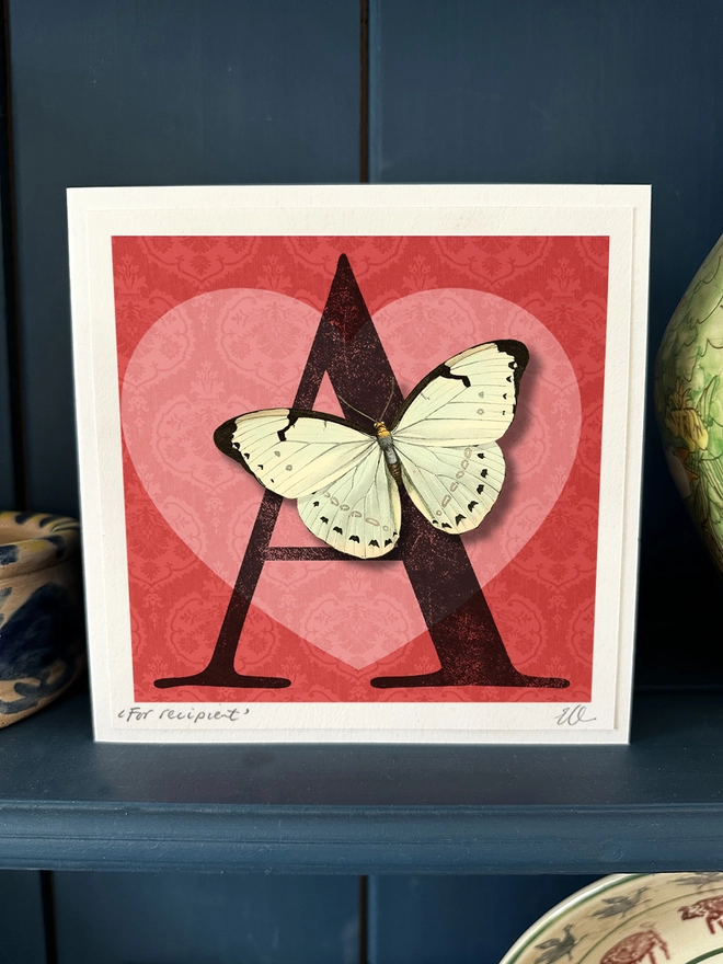 Valentines day card, showing a letter A which shows it is for pesonalisaion. The letter sits against a red background with a pink heart. Sitting on the letter A is a white butterfly. The butterfly is 3 dimensional and the wings can be folded open for display. The card is sitting on a blue dresser displayed in the home setting.