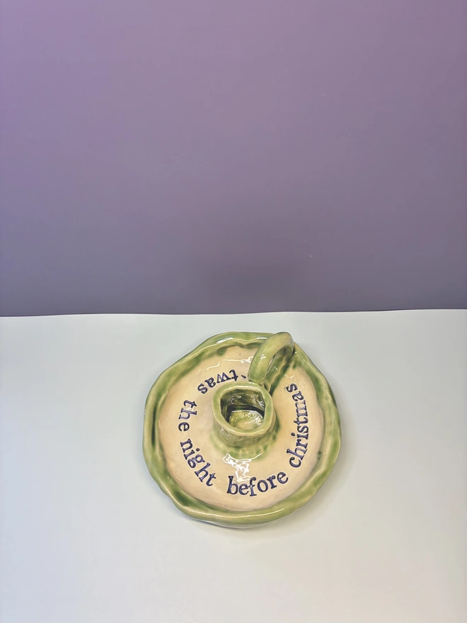 Handmade ceramic Christmas taper candlestick holder with a soft green glazed edge and the words “‘Twas the night before Christmas” in dark blue lettering, arranged around the candle holder opening. Photographed against a pale background.