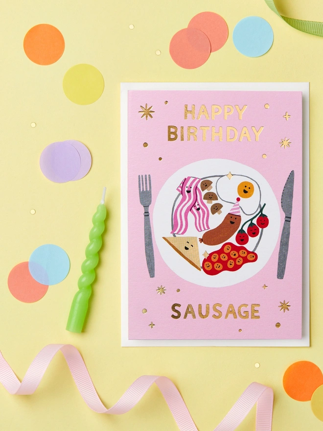 Happy Birthday Sausage card paired with a white envelope on a yellow background with confetti and streamers scattered around.