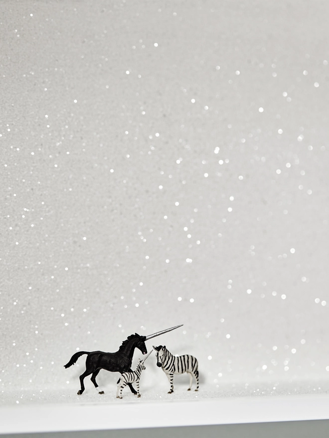 Miniature scene in an artbox showing a trio of tiny creatures - a black unicorn, a zebra and a baby ‘zunicorn’ (a mix of the zebra and unicorn) standing against a sparkling white backdrop 