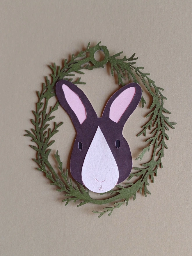 Black Bunny Easter Tree Decoration. Dark brown bunny face with white nose patch, black eyes and pale pink ears inside olive green foliage wreath oval