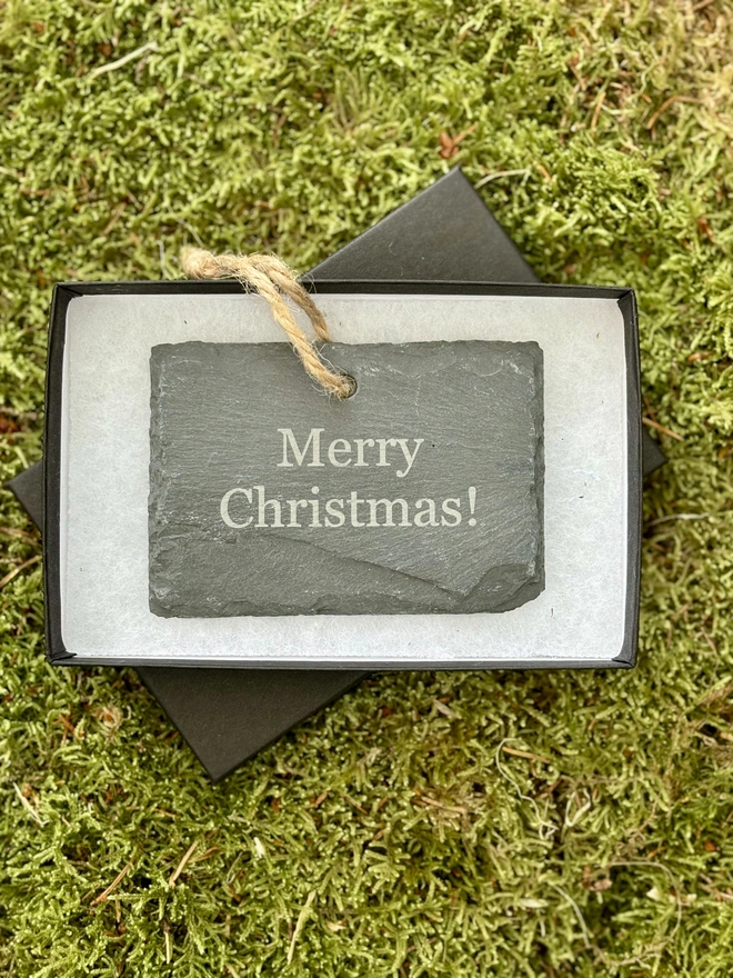 Hanging slate label personalised with Merry christmas in a black gift box on a bed of moss