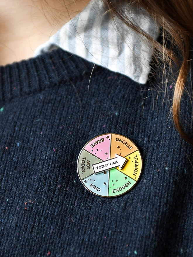 A round enamel pin that looks like a pie chart with six segments, each with positive word, and a white arrow that reads "Today I Am", is pinned to a navy blue jumper. 
