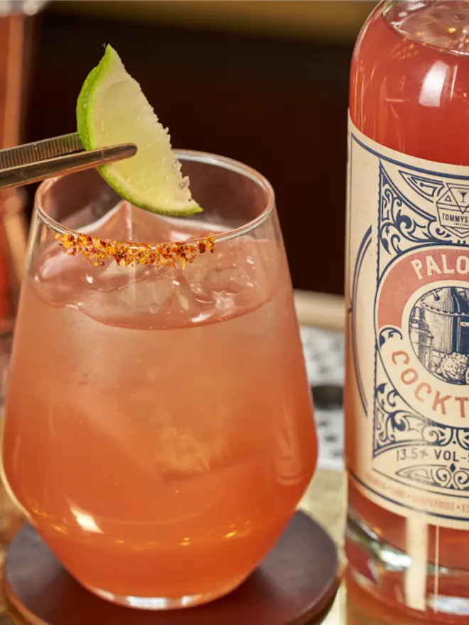 Tommy's Fizz Paloma Cocktail, a pink Paloma cocktail with a chili-salt rim and a lime wedge next to its bottle.