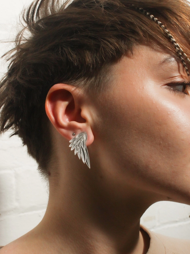 A woman earring Wing shaped Earrings in a Silver Acrylics  with a camel coloured jumper.