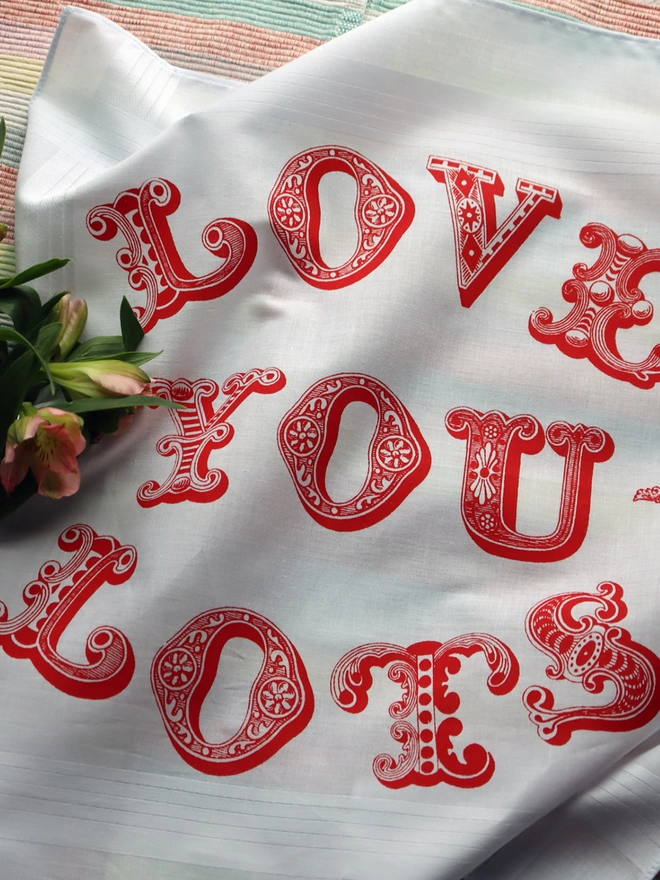 A Mr.PS Love you Lots hankie printed in red laid flat on a striped tablecloth with some fresh flowers