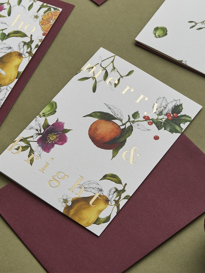 ivory based christmas card with festive hand drawn botanicals with gold foil wording and a burgundy envelope