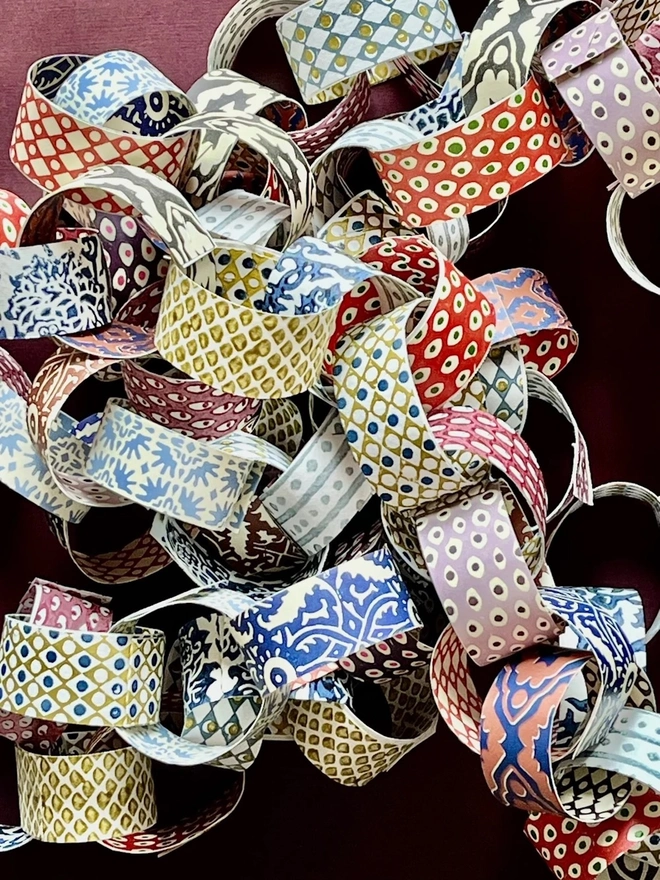 Colourful Paperchains (Set Of 60). A heap of linked paper chains in a mix of lattice, polka dot and floral patterns laid on a deep burgundy background. The multicoloured garland shows the full range of printed paper designs in shades of gold, blue, red and lilac.