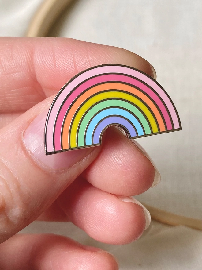 A small pastel rainbow enamel pin badge is being held.