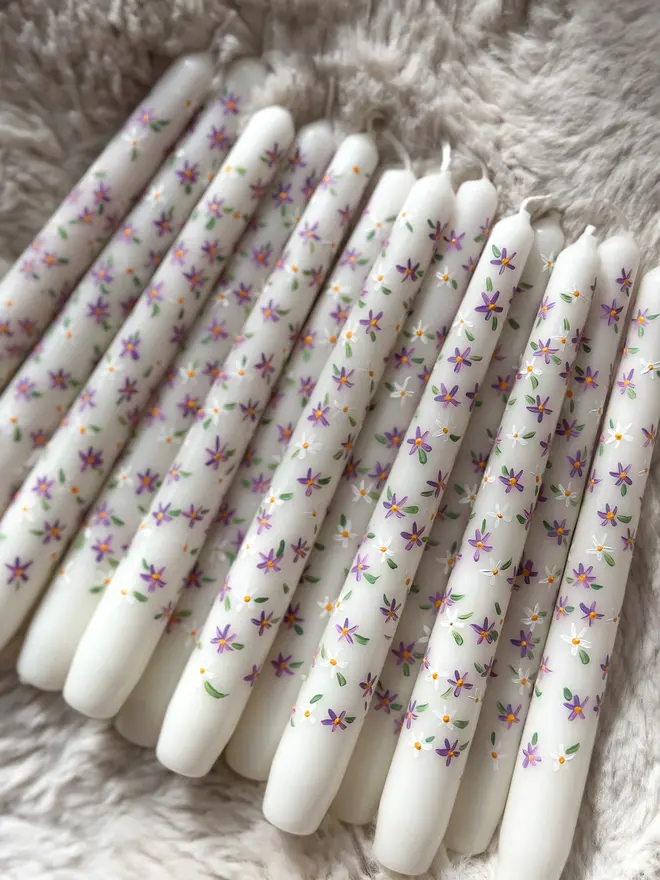 Purple Fleur Candle, a group of purple fleur candles sitting on a white rug. Their purple floral design can be seen wrapping around them. 