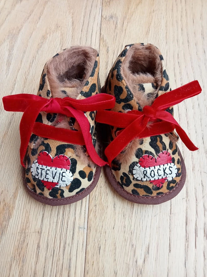 A pair of leopard print baby booties, lined with fur and with red velvet ribbon laces. A red heart with a white scroll adorns each booty. One says STEVIE, the other, ROCKS in black stitches