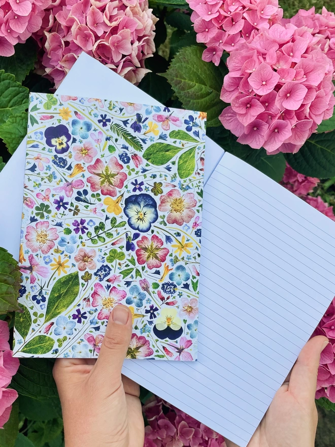 Hand Holding Pressed Floral Design Recycled Notebooks, One Open Showing Lined Pages, Pink Flower Background