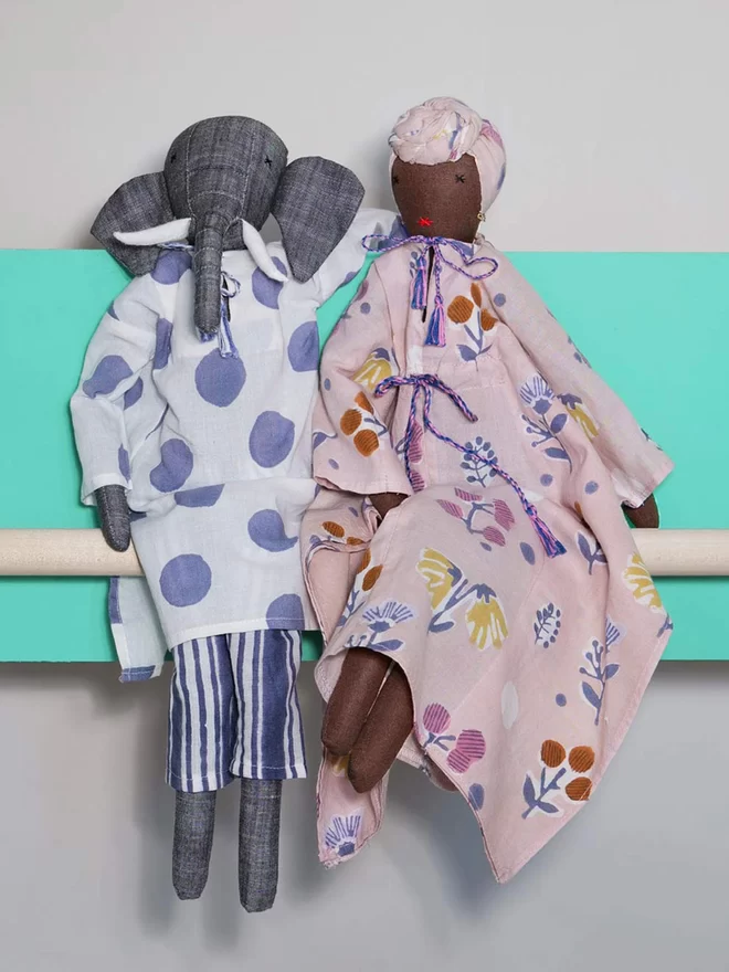 Elephant doll and black doll dressed in block printed outfits, one navy and white and one pink with multicoloured pattern, sitting on a wooden pole in front of a bright green wall