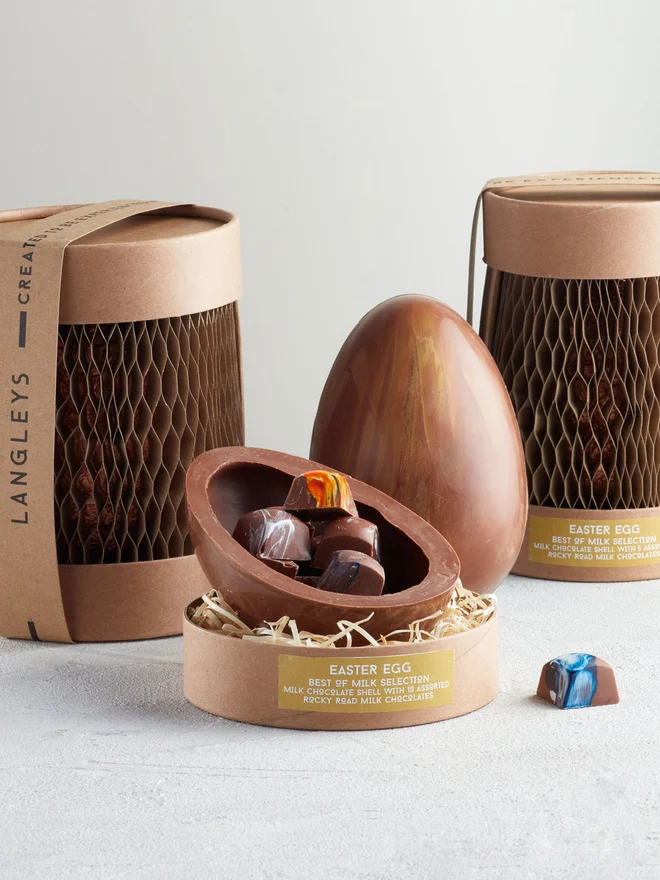 Best Of Milk Chocolate Easter Egg, a chocolate egg sitting on a white surface. It is accompanied by another chocolate egg that is opened showing the chocolates within. 