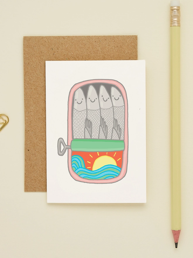 Close-up of mini sardines card on cream background with pencil and gold paperclips. Illustration shows four smiling fish in a tin with ocean waves and a rising sun.