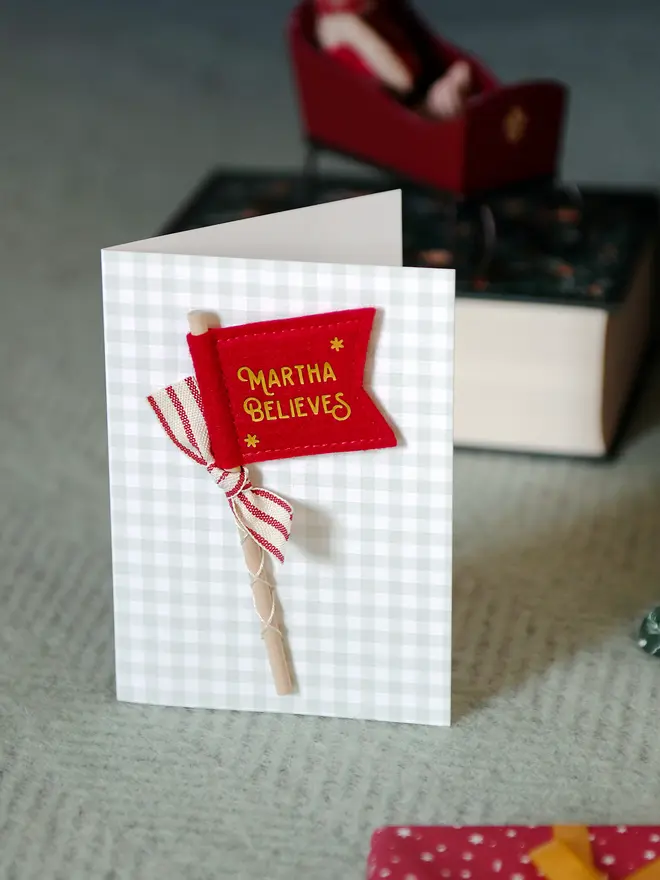 A Christmas card with a tiny handmade felt flag attached. The flag reads 'Martha Believes' and has a red striped ribbon tied to it.