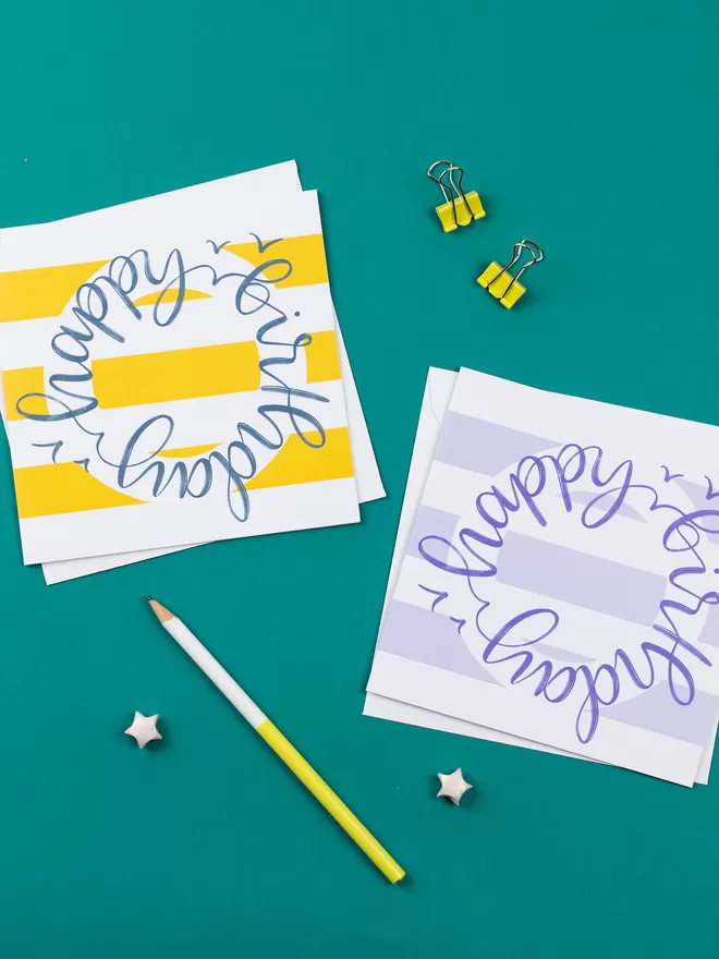 Two colourful stripy birthday cards lie on a teal background, surrounded by stationery items