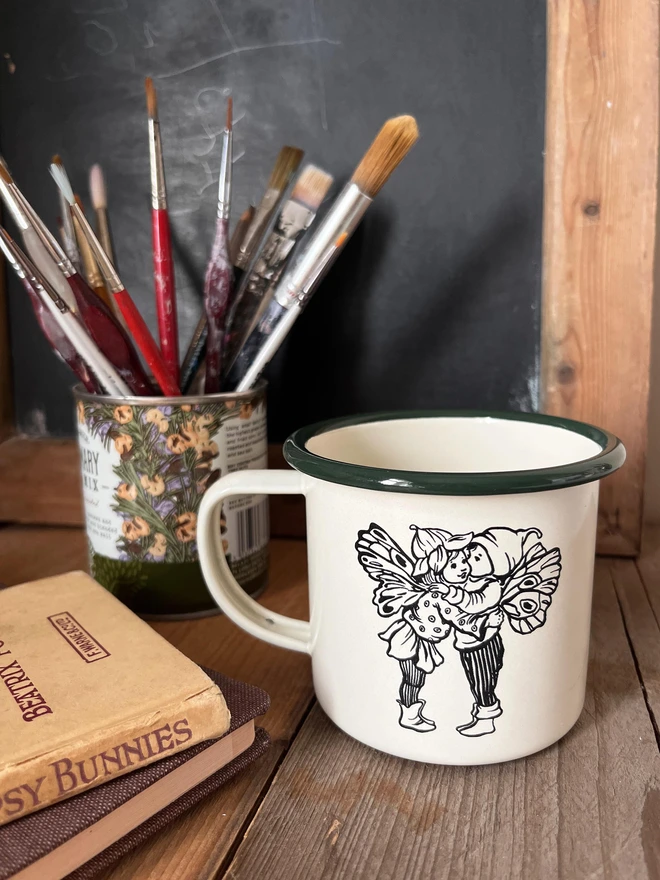 a cream enamel mug with a bottle green rim etched with an illustration of 2 cute pixies embracing