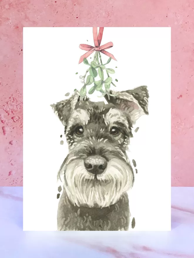 A Christmas card featuring a hand painted design of a Schnauzer, stood upright on a marble surface.
