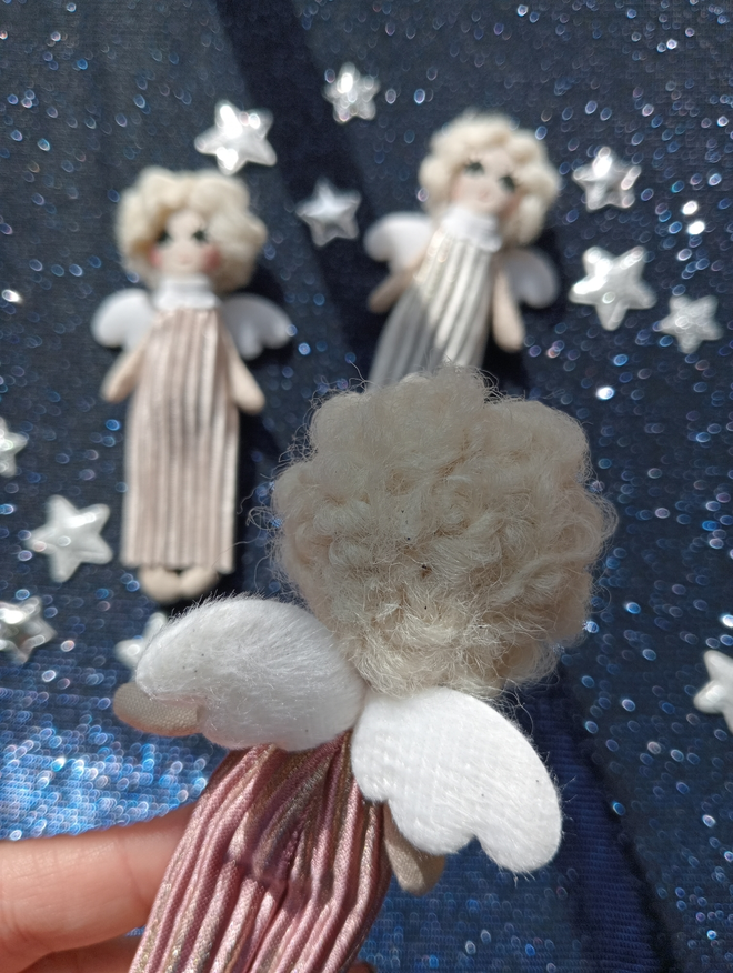 the back of a tiny angel doll in the foreground. behind her are two more tiny angel dolls on a dark blue glittery background surrounded by stars