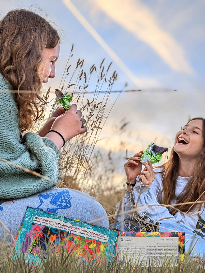 two happy girls in a field enjoying charity white chocolate wrapped in green foil & floral packaging