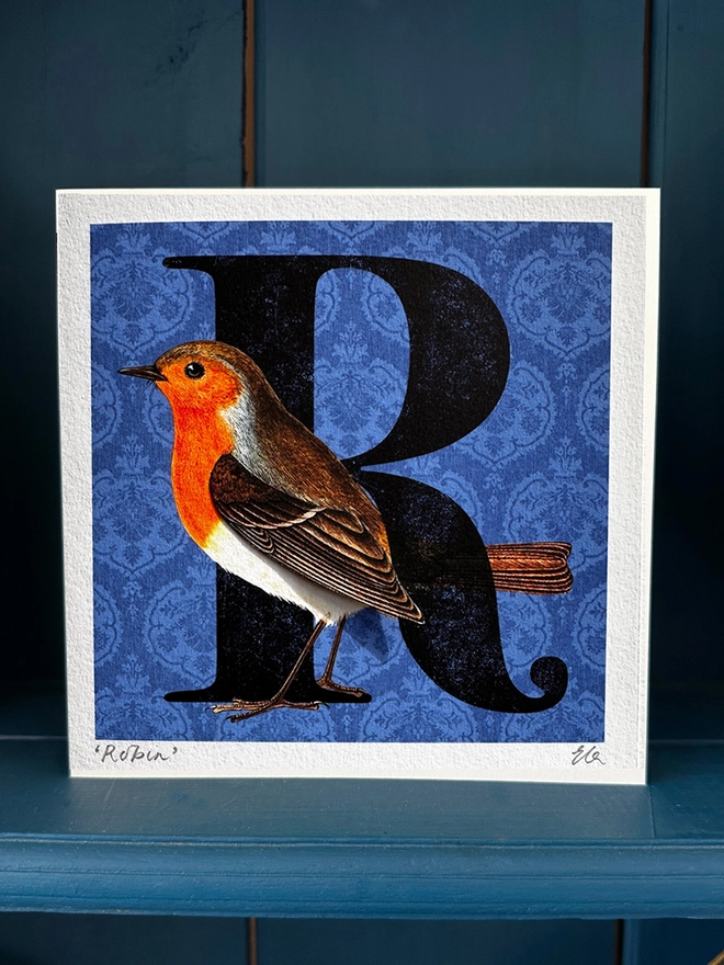 Photo of a handmade card displayed on a blue dresser showing a Robin on a blue background with a big letter R.