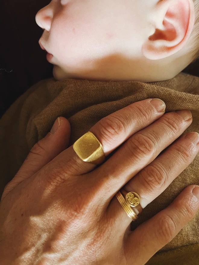 Image of a Hand wearing 3 gold rings, one square, one band and one gold moon face ring. Hand resting on the sleeping chest of a baby.
