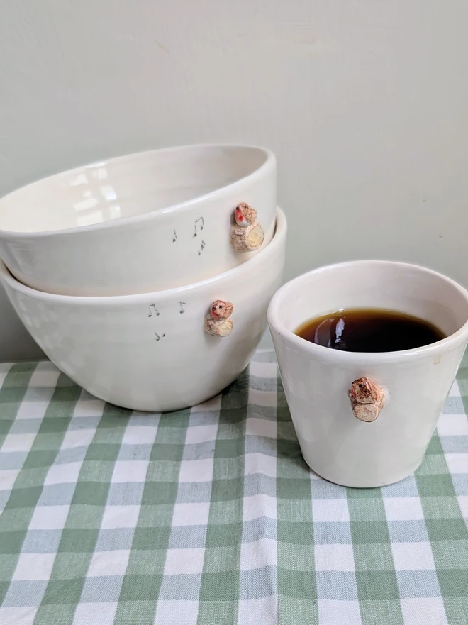 stack of two white ceramic bowls each with a tiny modelled robin perching on a modelled branch with musical notes resting on a green gingham tablecloth with a white espresso robin bird cup next to them