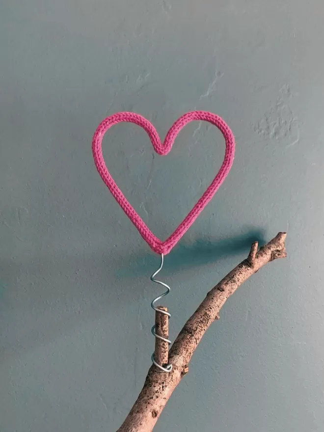 A pink string wrapped wire heart shaped Christmas tree topper on a branch against a grey background
