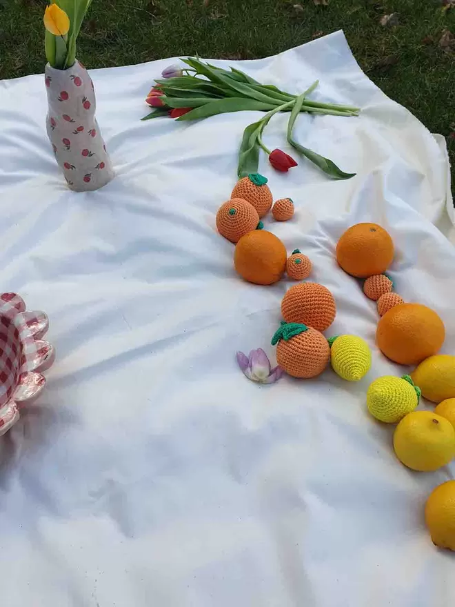 Crochet and real lemons and oranges on a white fabric background with tulips and handmade ceramics in the background. 