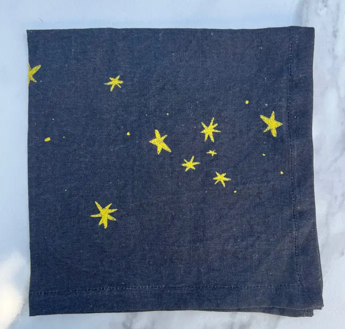 Stars Napkins In White (Set of 4). White cloth napkins with scattered yellow star prints, placed on a Stars Napkins In White (Set of 4). White cloth napkins with scattered yellow star prints, placed on blue table cloth. 