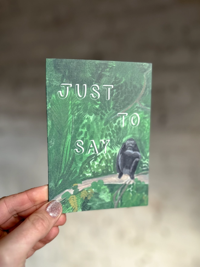 a greetings card featuring an illustration of a chimp sitting in the jungle with the text “just to say”