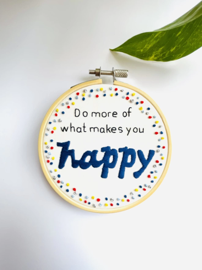 Hand embroidered hoop wall hanging reads Do more of what makes you happy in black and blue stitching with a colourful french knot border