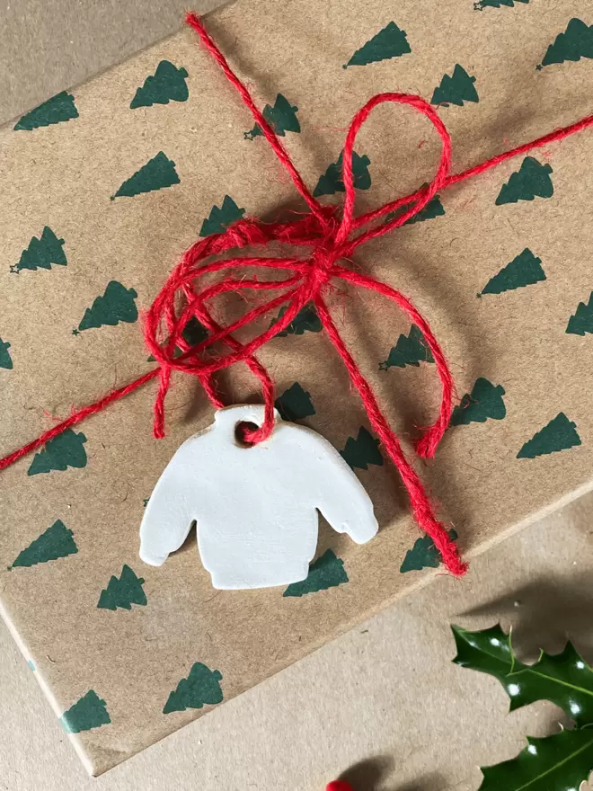A small clay white jumper tag attached to a present with red twine. The present is in a brown box with green christmas trees on.
