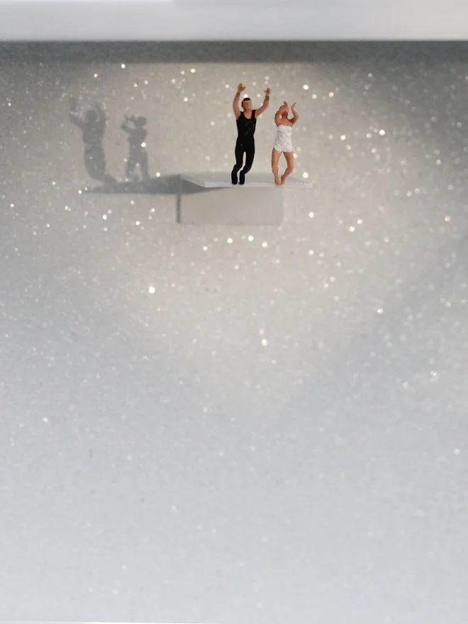 Miniature scene in an artbox showing a tiny woman and man about to dive off a diving board, set against a sparkling white backdrop (close up). 