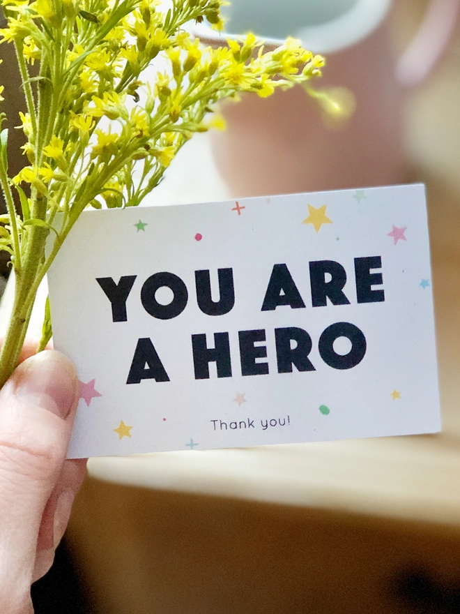 A mini thank you card with the words "You are a hero" and a gentle star design is being held beside a bunch of yellow flowers.