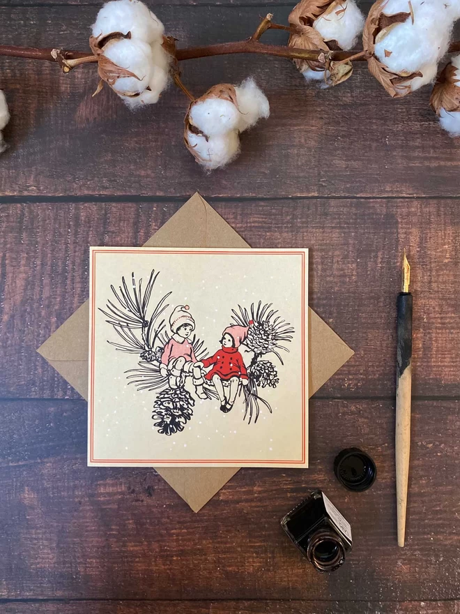 a Christmas card featuring a drawing in ink of 2 pixie children sat on a pinecone branch