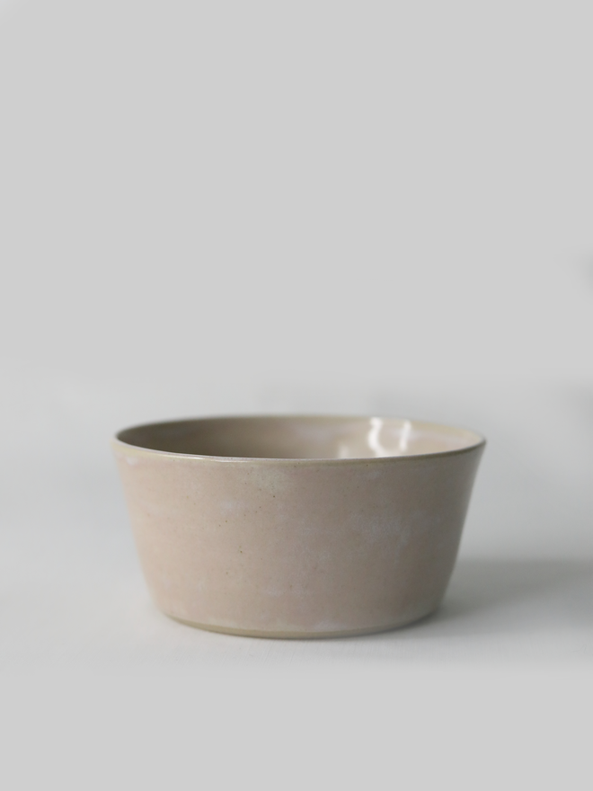 A single hand-thrown ceramic breakfast bowl in blush pink, with a glossy glaze showing subtle variation in tone, a gently flared form and unglazed natural clay base, photographed against a plain light grey background.