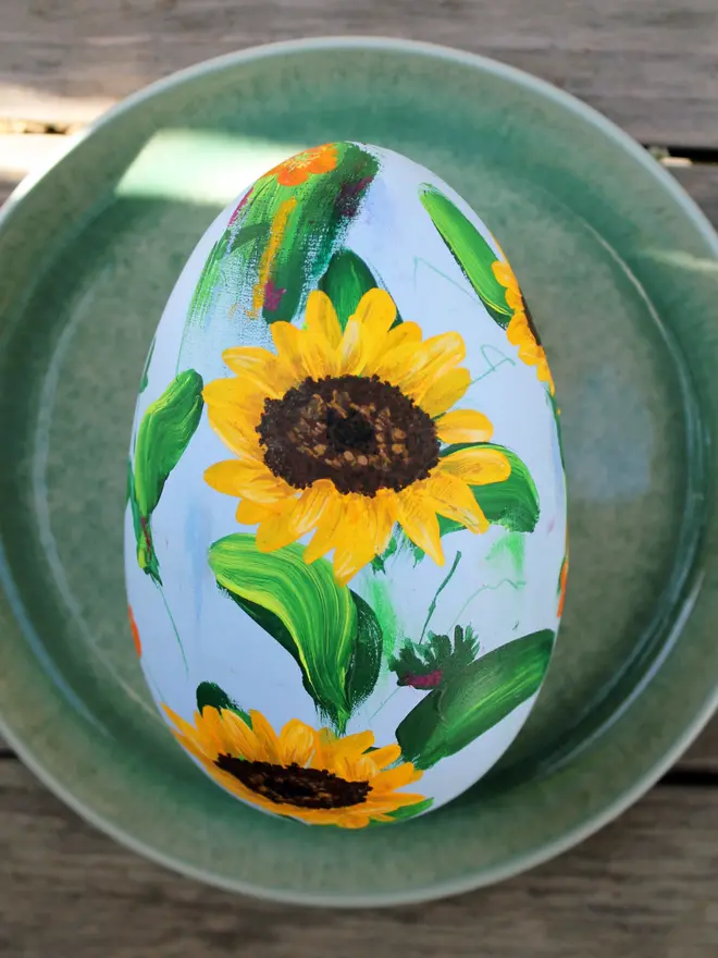 pale blue floral hand painted wooden egg, adorned in sunflowers and orange Icelandic poppy flowers, positioned on a green plate