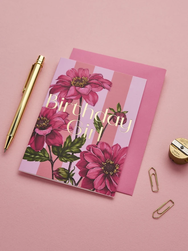 'Birthday Girl' card with gold foil wording and illustrated Dahlia flower on a pink and lilac striped base and a hot pink envelope
