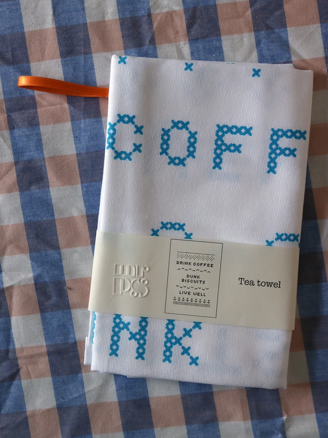 A Mr.PS folded tea towel on a gingham tablecloth