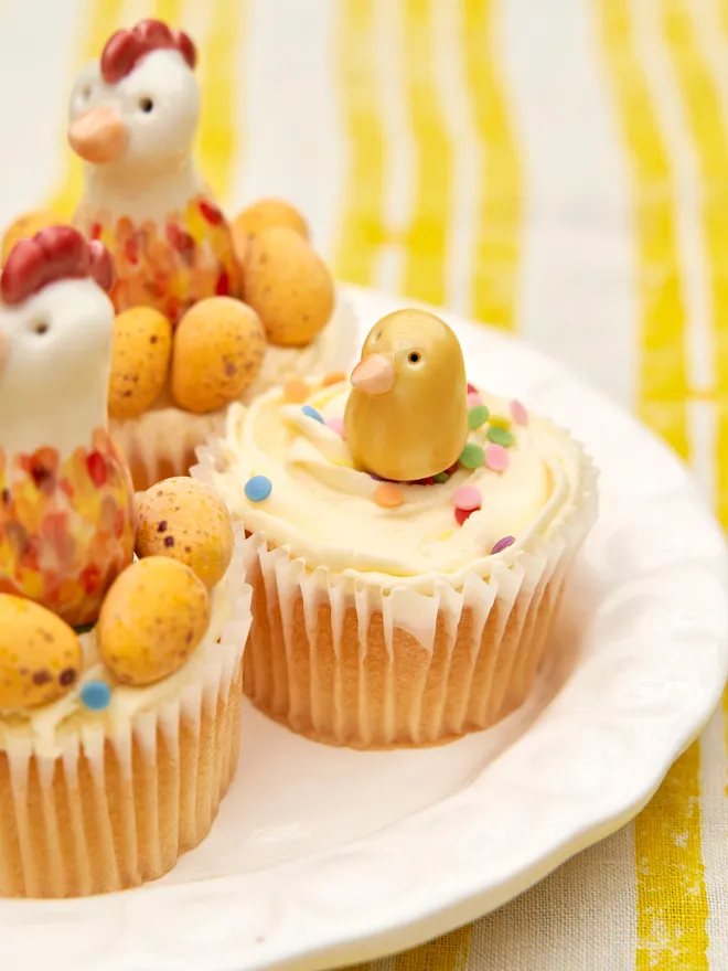 2 ceramic Easter chicks and 2 ceramic Easter chickens on top of 4 cupcakes with mini eggs and sprinkles, on a plain white plate on a stripy yellow and white tablecloth