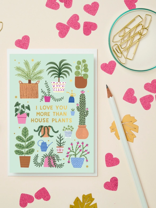 I Love You More Than  House Plants illustrated card paired with a white envelope on a cream background with heart-shaped confetti and stationery around it.