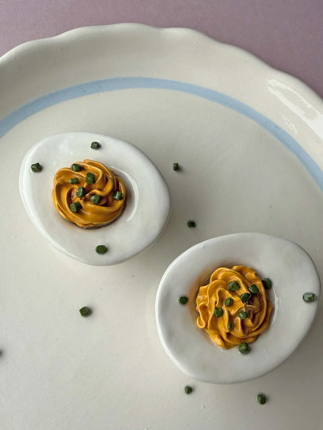 Ceramic devilled eggs with chive detail, on a plain white plate with blue trim, on a pink background