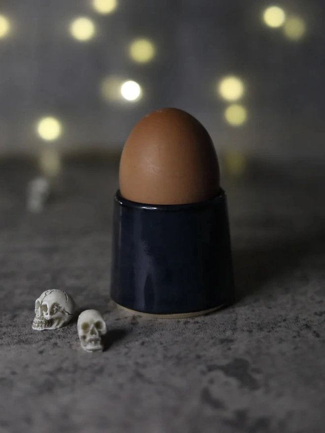Classic Handmade Egg Cup, a ceramic egg cup sitting on a dark surface board against a fairy light backdrop. It is accompanied by some mini skulls. 