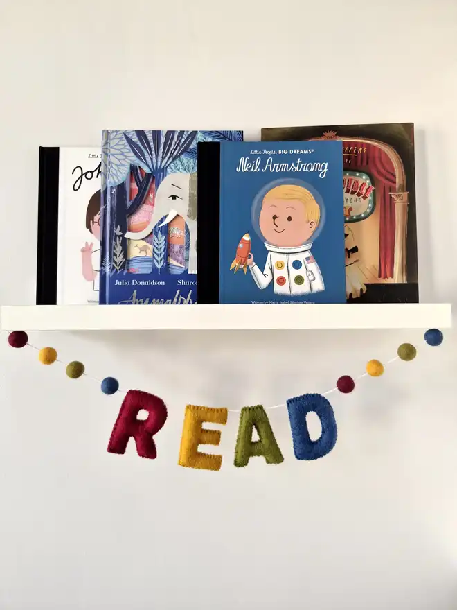 A dark rainbow felt banner hanging under a shelf full of children's book. The garland says 'Read' with felt balls either side.