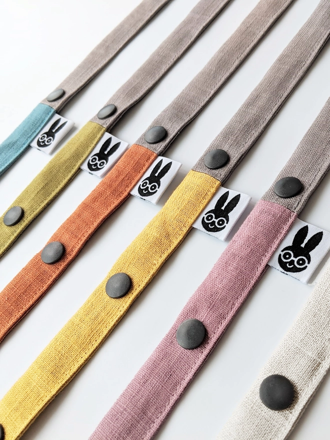 Handmade toy straps in soft neutral and pastel tones, featuring sturdy snap fastenings and small fabric tags with a bunny logo, neatly arranged in a row.