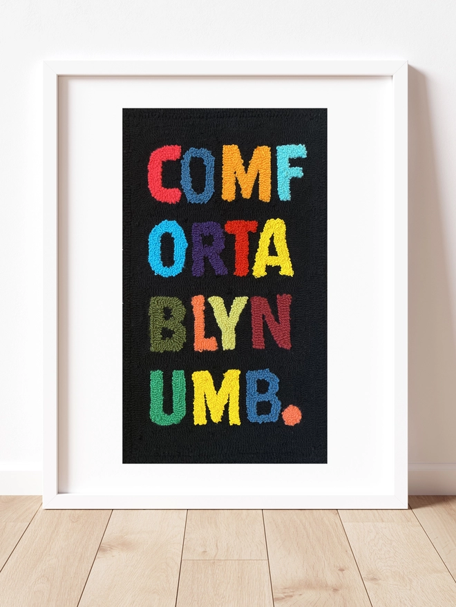 Comfortably numb in bold primary colour upper case letters on a black wool background framed in a white box frame stood on a wooden floor