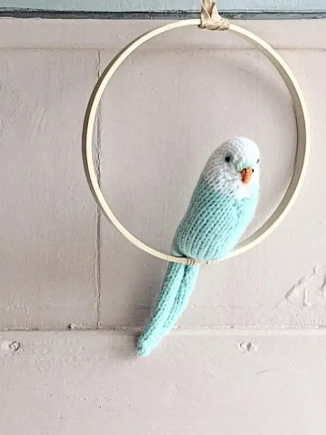 Colourful Hand Knitted Budgerigar, a blue colourful birds sitting in a wooden ring against a white backdrop. 