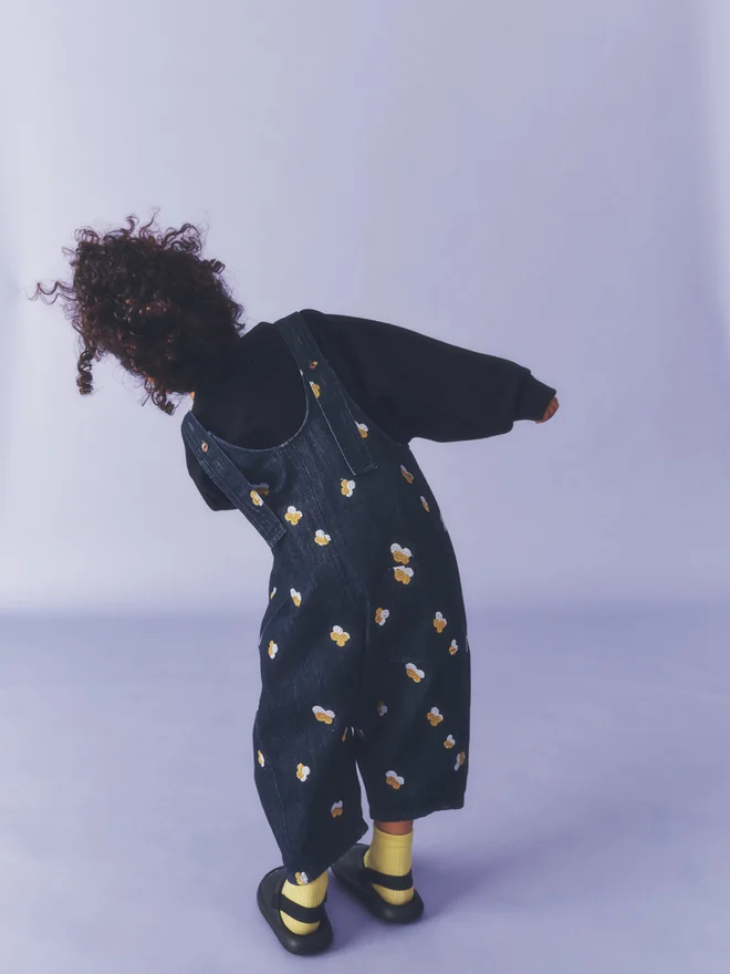 Back view of a child leaning forward against a lilac backdrop, showing the embroidery and patch pocket of Studio Koter Kai dungarees in Denim Petal, worn over a black sweatshirt.